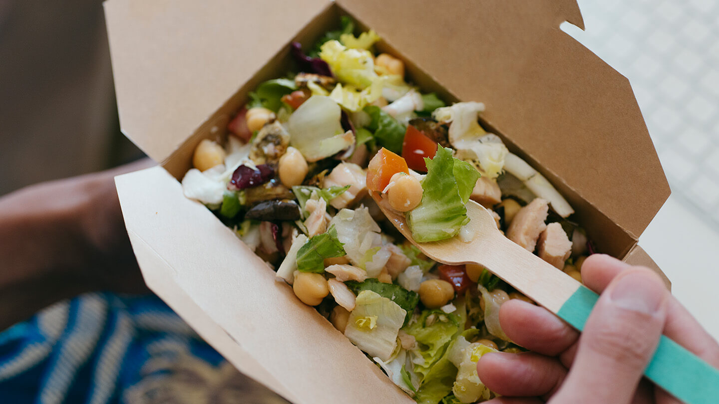 top view of a salad in a takeout container being eaten by an unseen consumer with a sustainable wooden fork